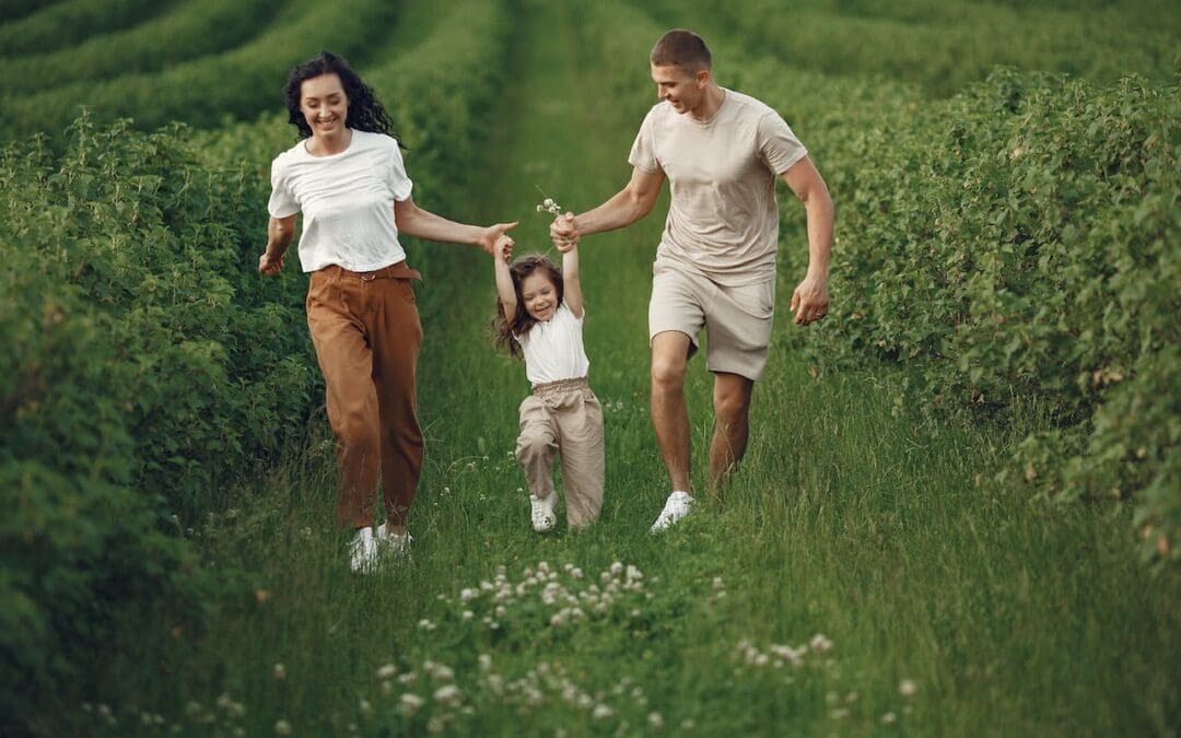 A smiling, young white couple run through rows of green plants, each holding the hand of a young girl as she swings between them and clutches white flowers.
